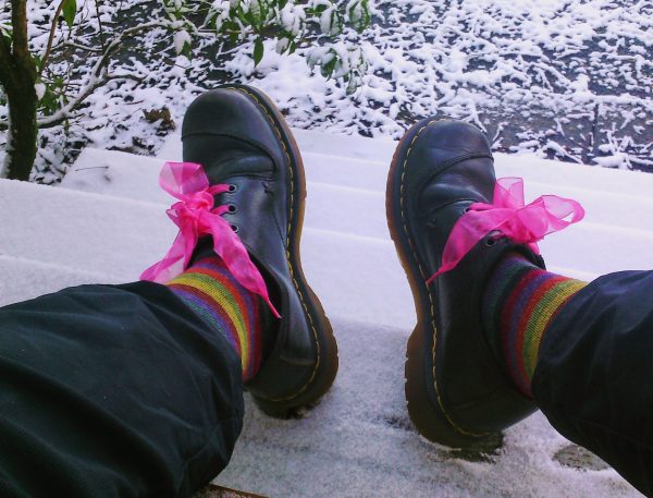 feet wearing doc martens with pink chiffon shoelaces, and stripey socks, against a snowy background