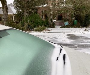 Car windshield coated in pebbly ice, with wipers covered by snow. Beyond is a snowy street and a house behind bare trees.