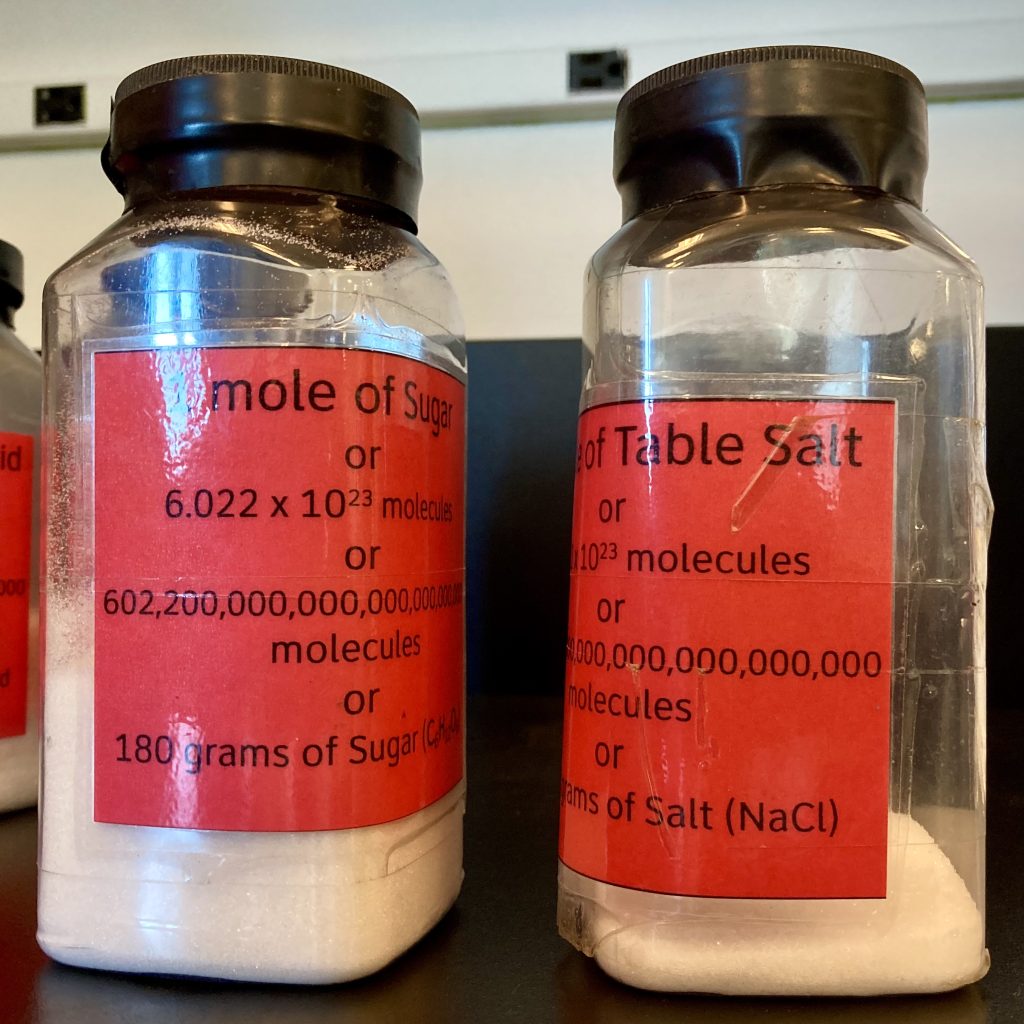 two labeled clear plastic bottles: one contains a mole of white sugar, the other contains a mole (much less volume) of table salt.