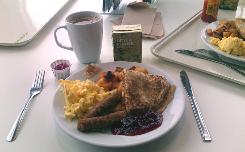 IKEA breakfast with eggs, sausage, pancakes, potatoes, and jam. Cup of coffee and juice box behind plate.