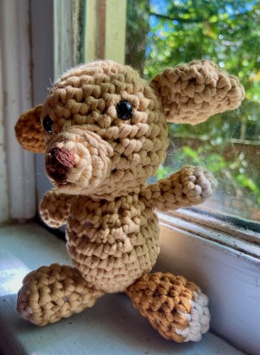 crocheted dog amigurumi, light brown and medium brown with embroidery-thread nose. The dog is a bit lumpy and haphazard, and sits on a sunny windowsill.