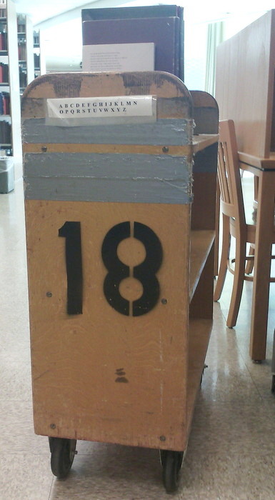 wooden library shelving cart with a stenciled 18 and a taped-on printout of the alphabet in capital letters