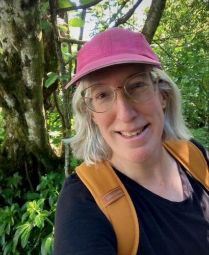 selfie of a white woman with a gray bob, glasses, and pink ball cap, wearing a backpack and standing in front of a hazelnut tree and other greenery