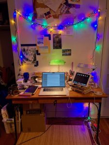 a wooden table lit with banker's lamp and string lights, piled with books, a laptop, pens, and cords.